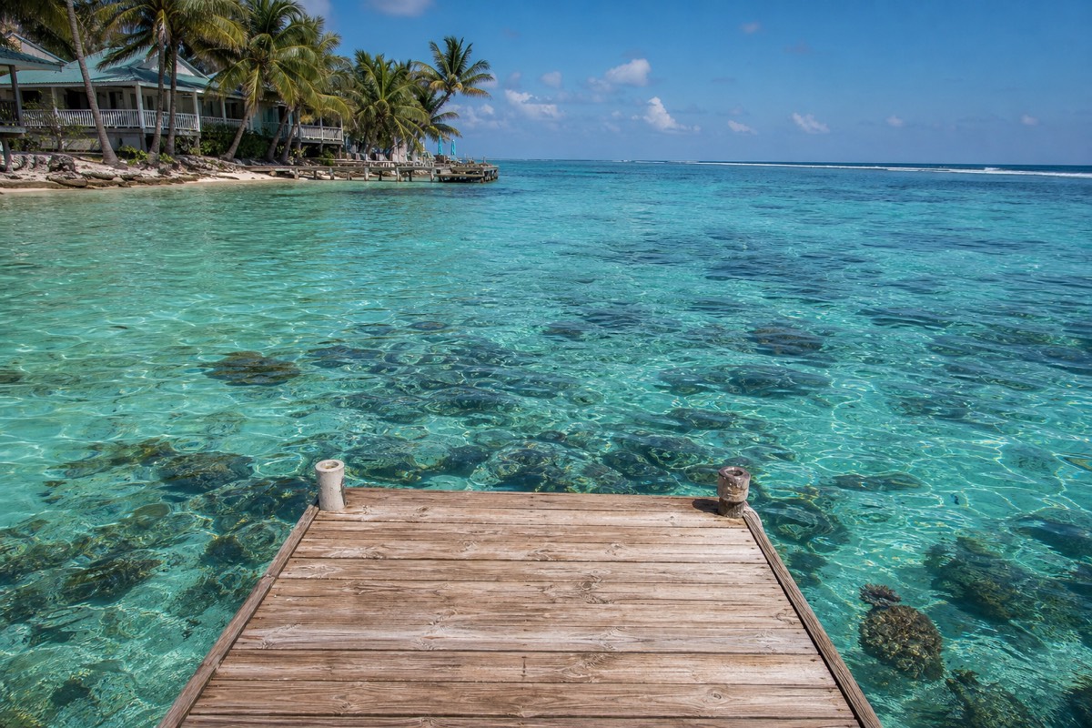 A dock reaching over shallow turquoise water on a Belize caye, with the barrier reef breaking in the distance